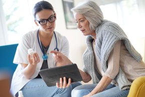 Kontrolle Zwei Damen, eine davon Ärztin, sitzen beisammen mit einem Tablet-PC und betrachten eine Dose mit Tabletten.